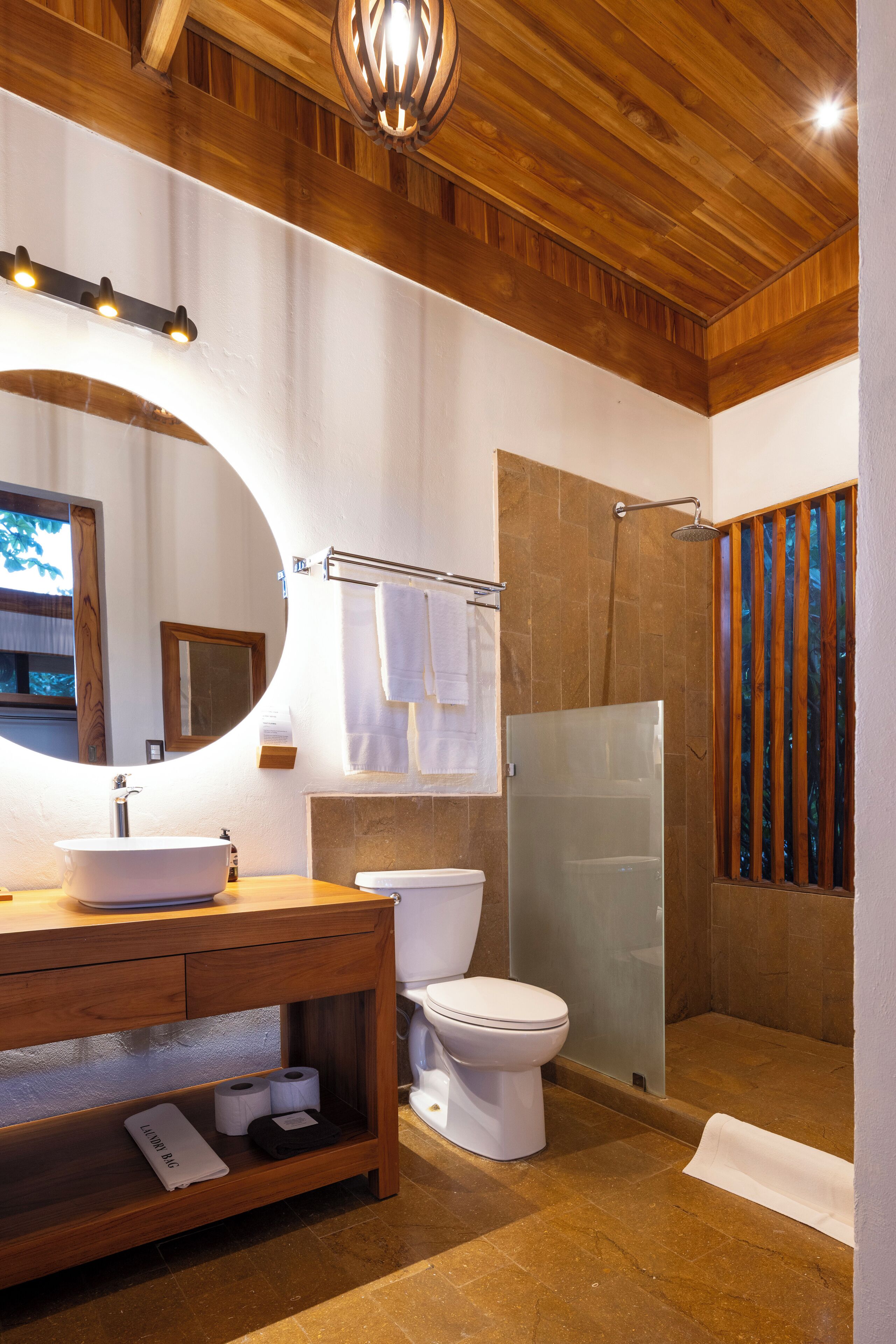 A bathroom with a wooden vanity, a round mirror, a toilet, a glass shower enclosure, and a towel rack.
