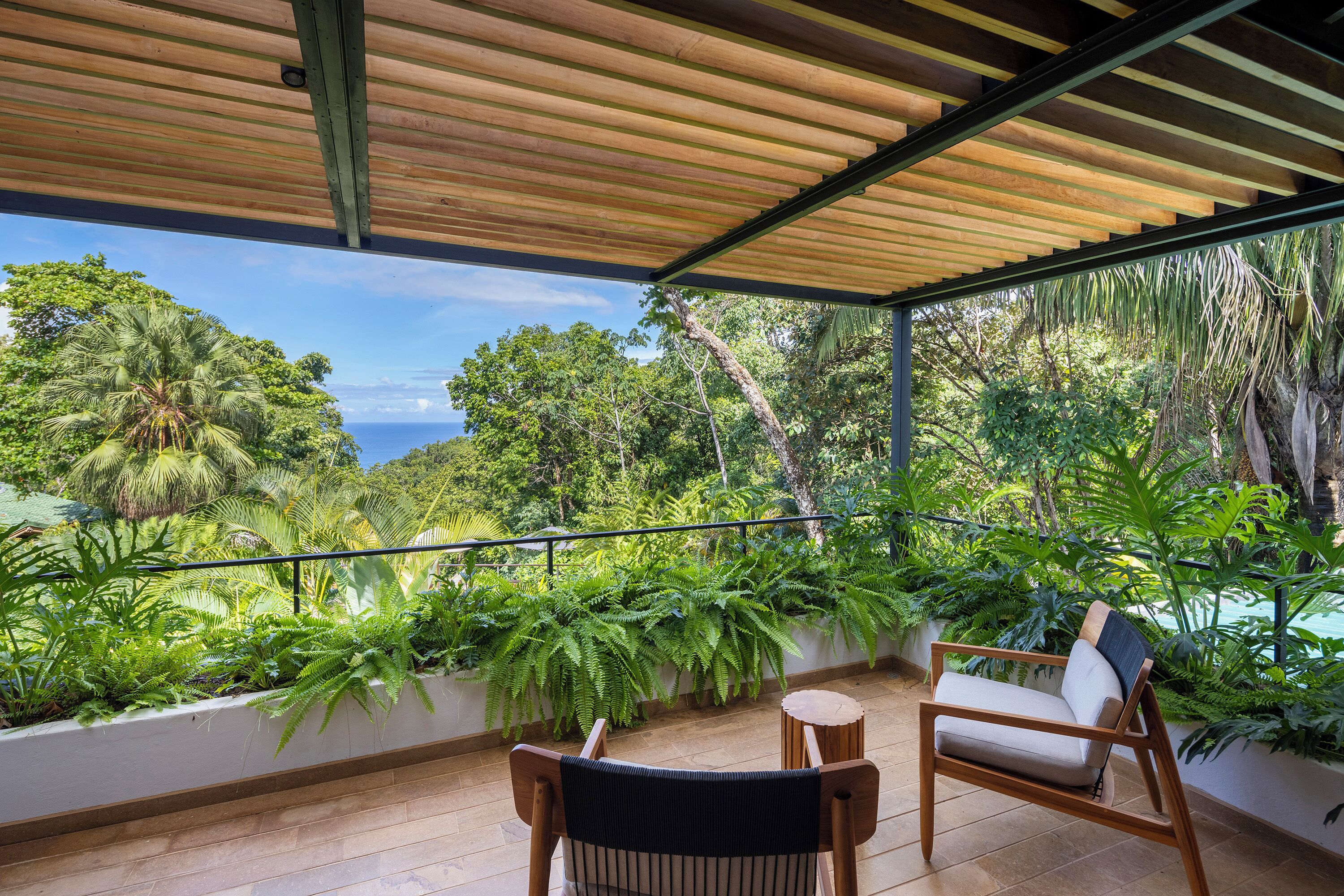 A covered balcony with wooden flooring, two wooden chairs, and lush greenery.