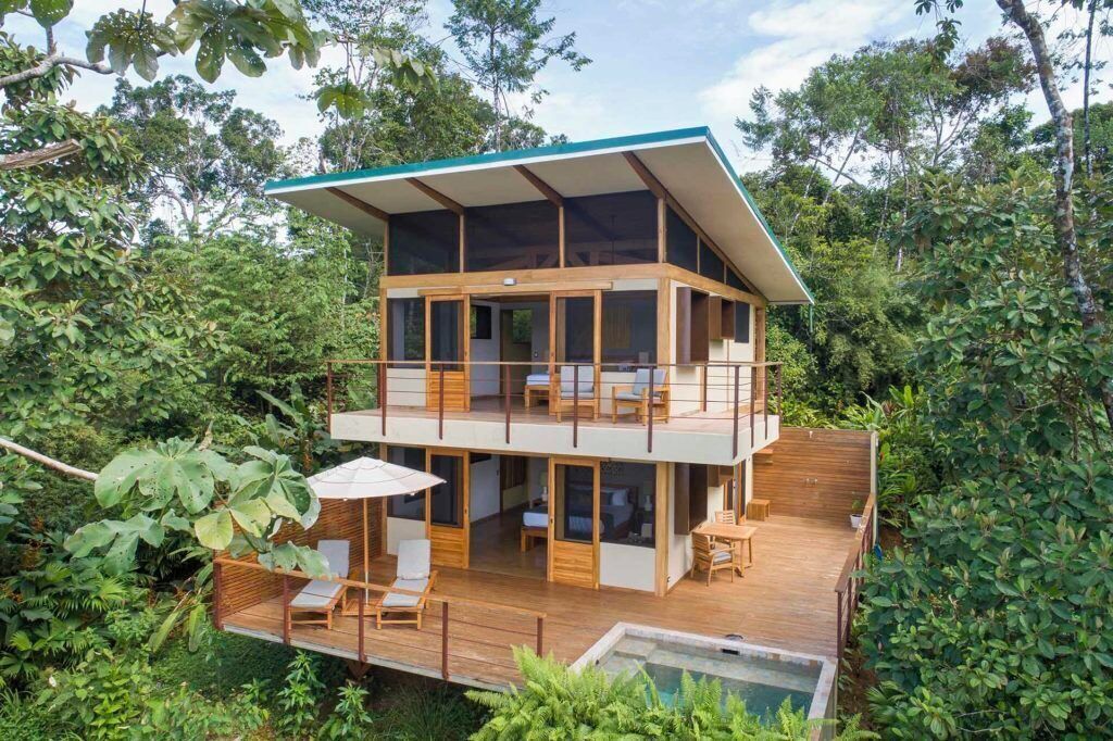 A two-story wooden house with a balcony, surrounded by lush greenery.