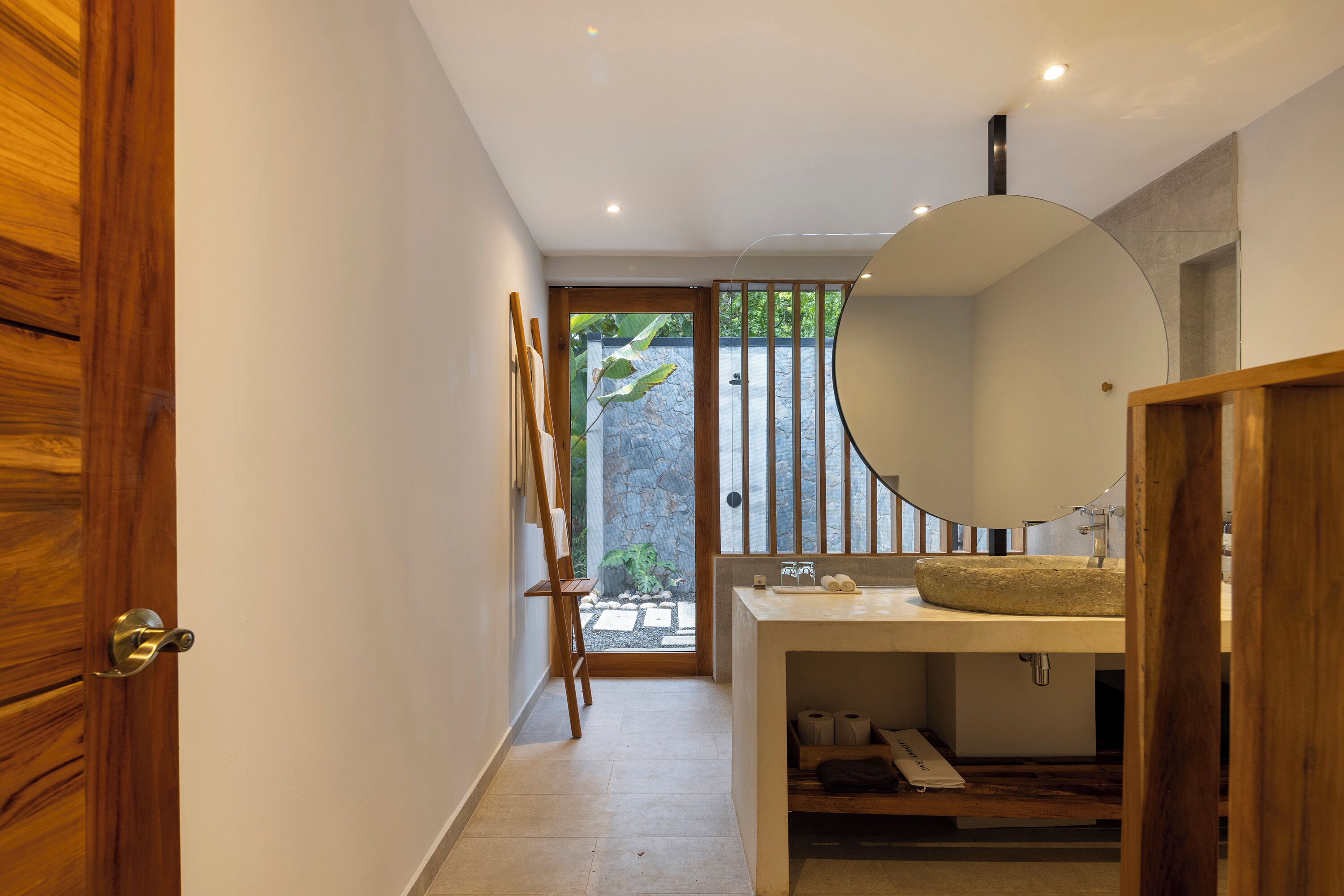A modern bathroom with a wooden door, a large round mirror, a stone wall, and a view of greenery outside.