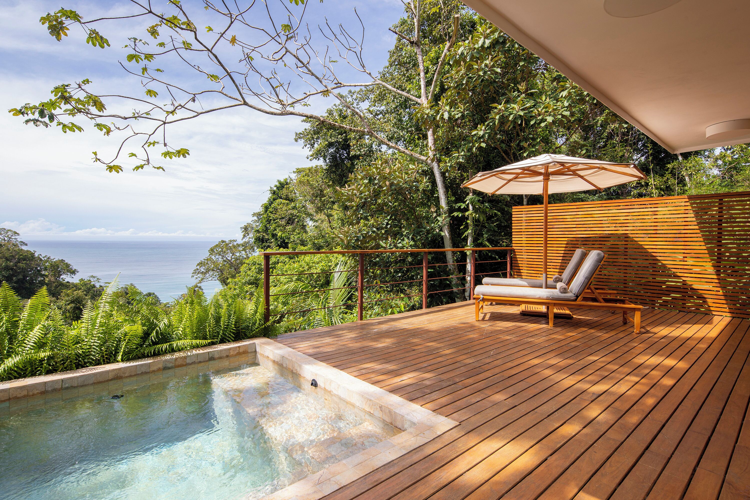 Une terrasse en bois avec un jacuzzi, une chaise longue et un parasol, offrant une vue sur un paysage luxuriant et la mer.