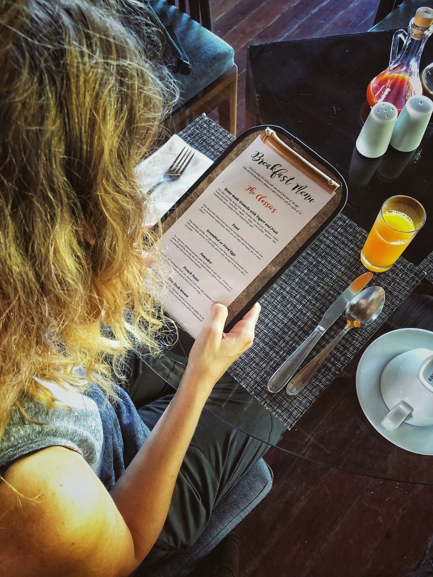 A person holding a breakfast menu at a table with a glass of orange juice, salt and pepper shakers, and a cup.