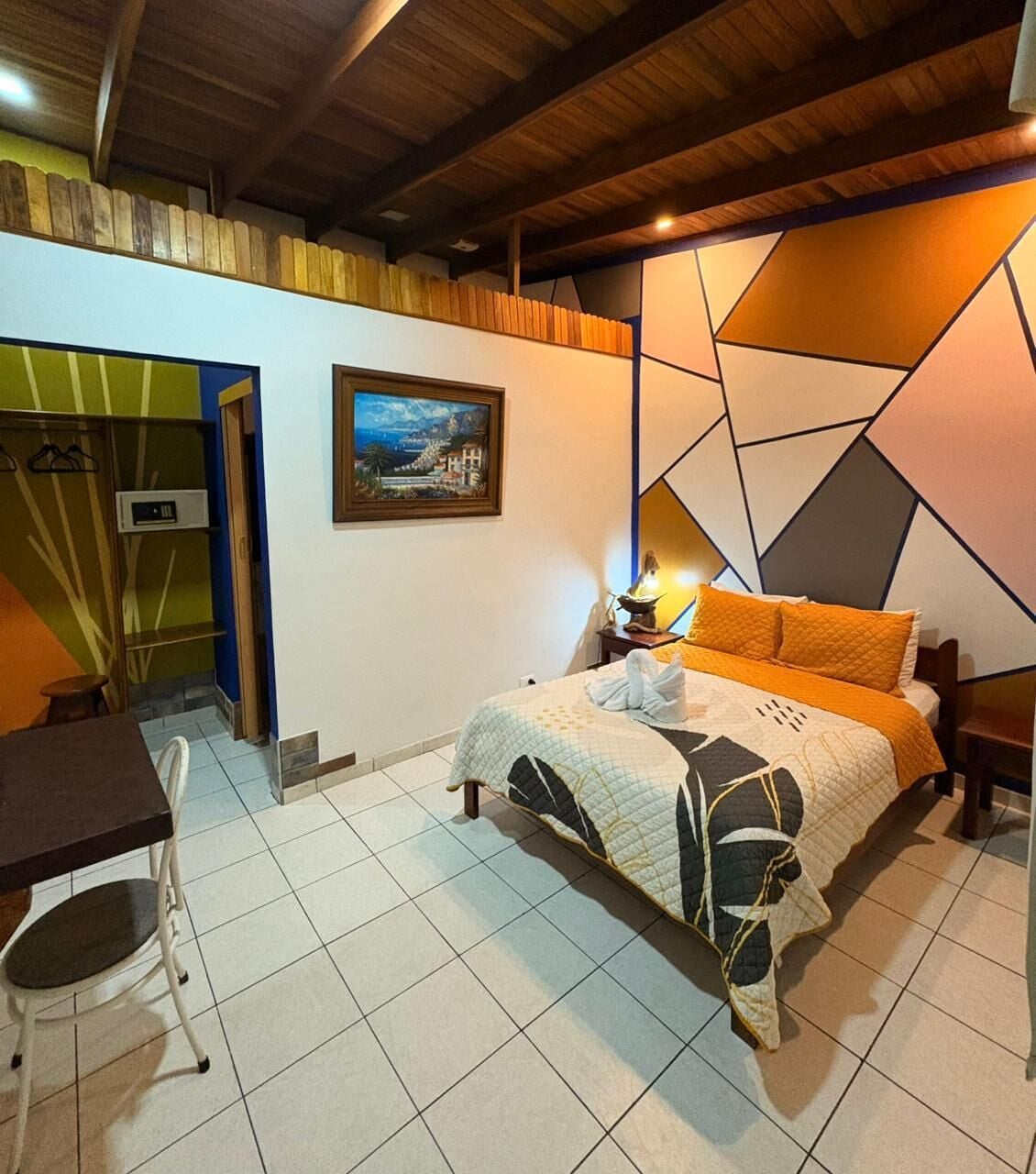 A bedroom with a bed, a desk, and a chair. There is a painting on the wall and a wooden ceiling.