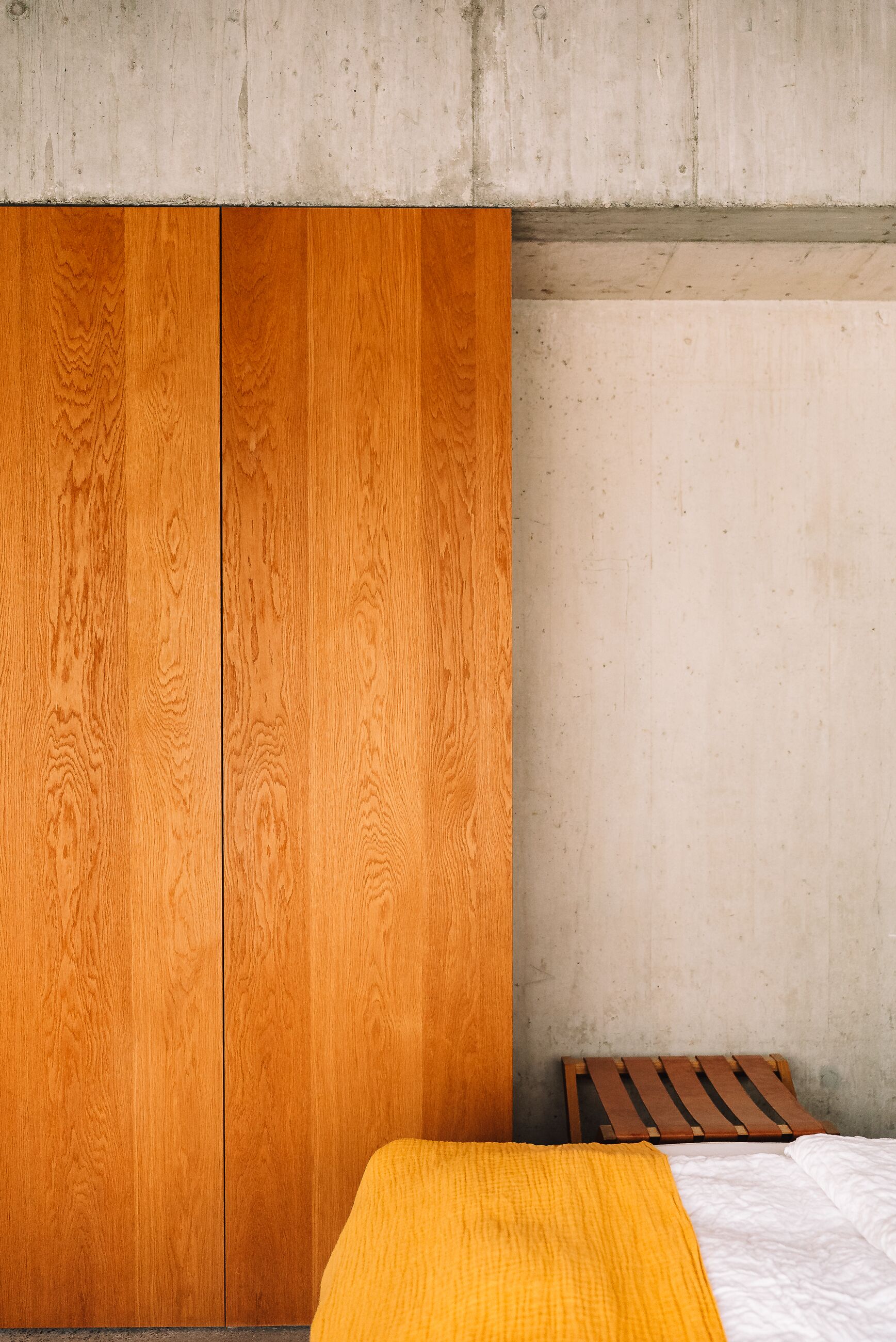 A bedroom with a wooden wardrobe, a bed with a striped headboard, and a concrete wall.