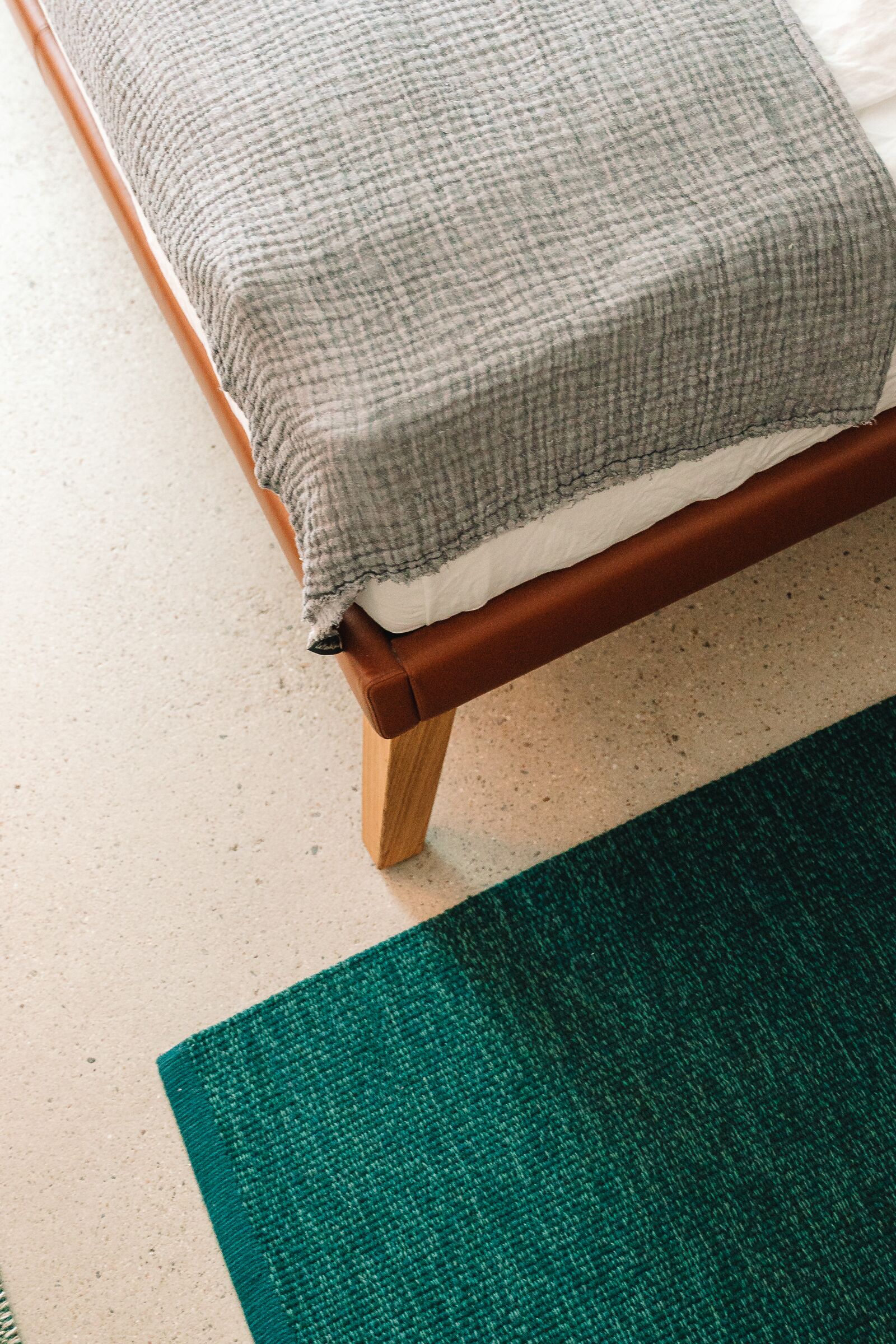 Una cama con una manta gris de cuadros, un cabecero de madera y un tapete verde sobre un suelo de tonos claros.