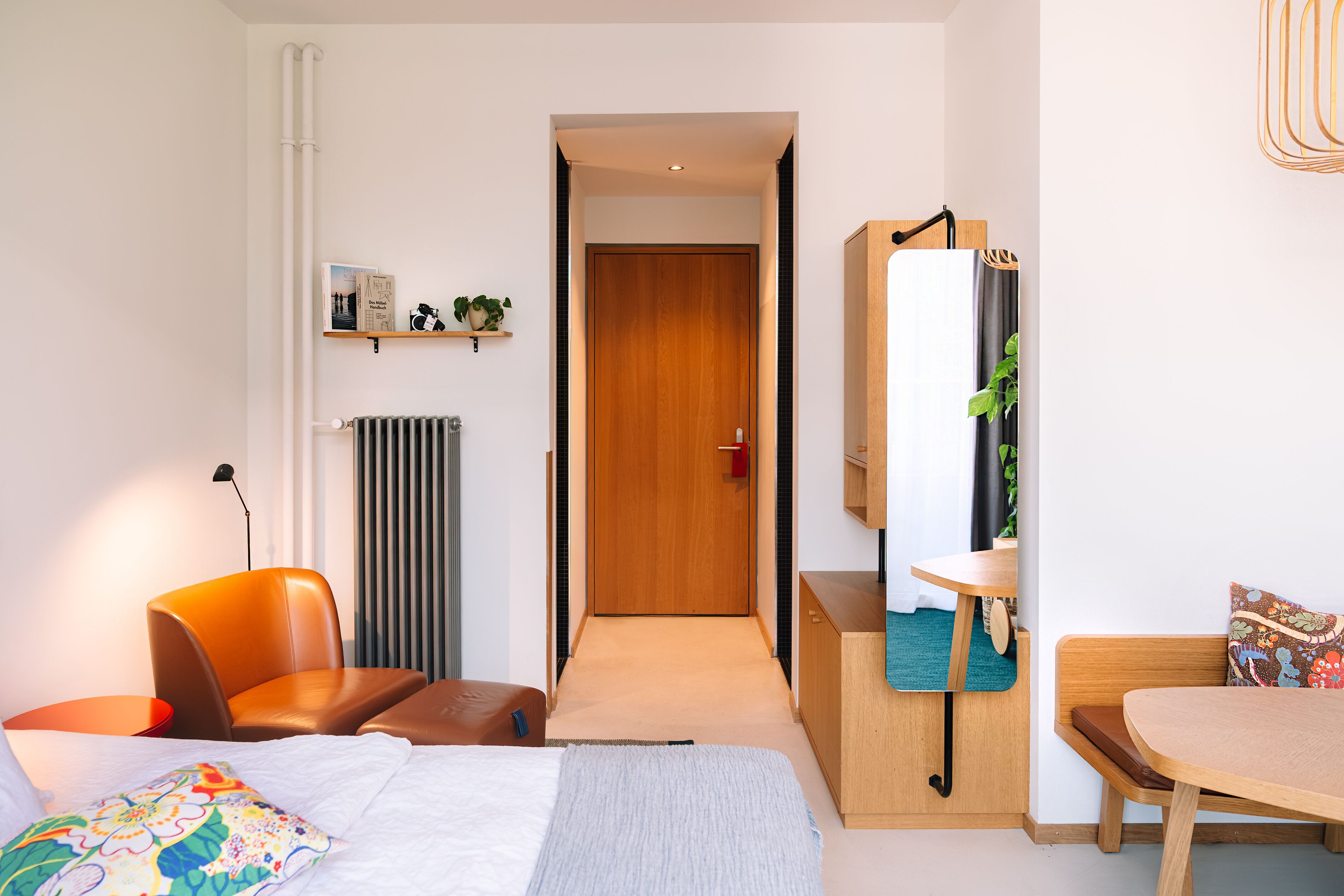 A hotel room with a bed, a chair, a table, a mirror, a shelf with a plant, and a door.