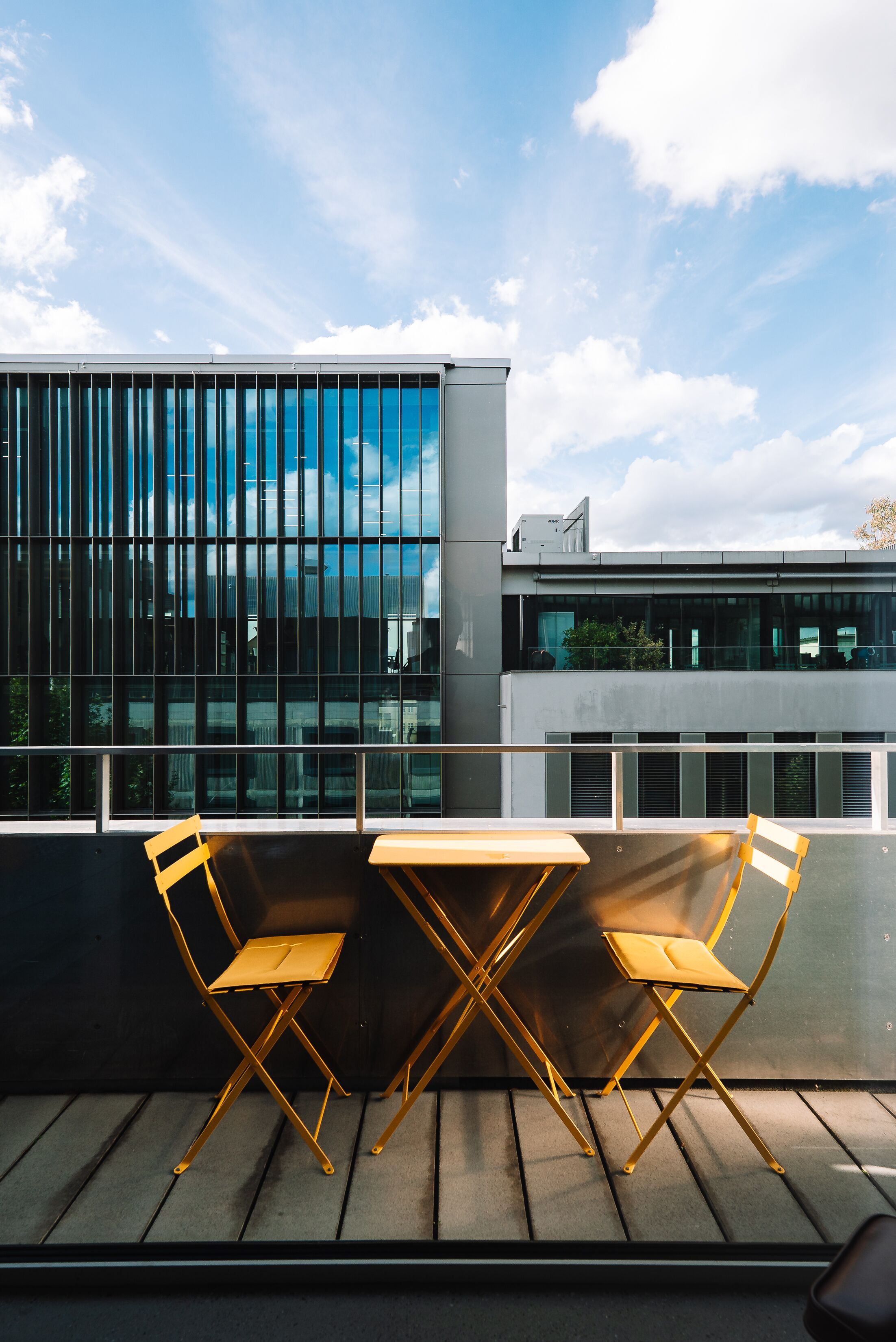 Un balcón con dos sillas plegables y una mesa, con vista a un edificio moderno con ventanas grandes y cielo despejado.