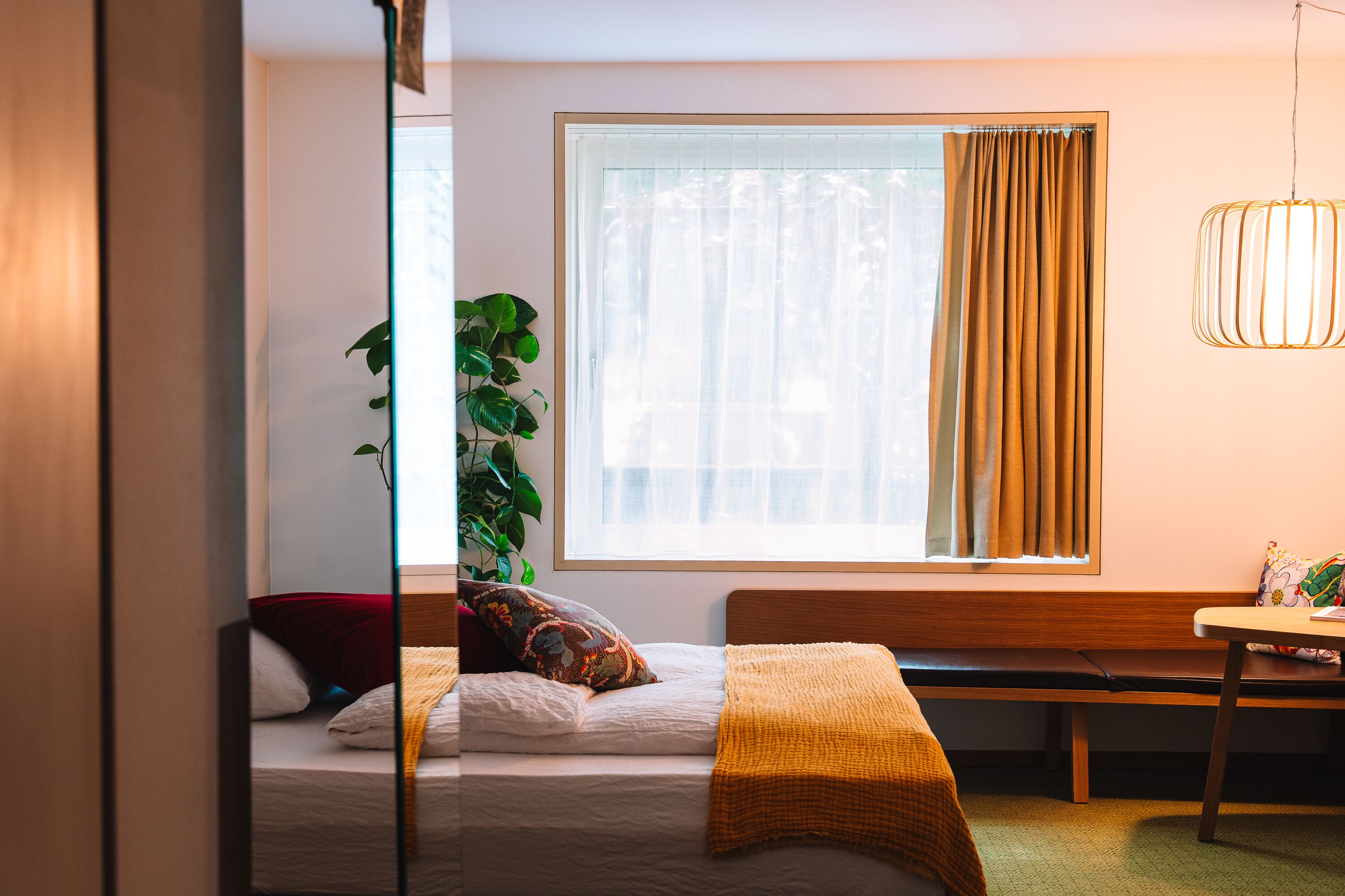 A bedroom with a bed, a bench, a table, a plant, and a window with curtains.