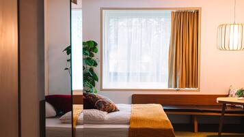 A bedroom with a bed, a bench, a table, a plant, and a window with curtains.
