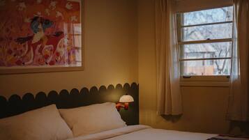 A bedroom with a bed, a nightstand with a lamp, a window with curtains, and a painting on the wall.