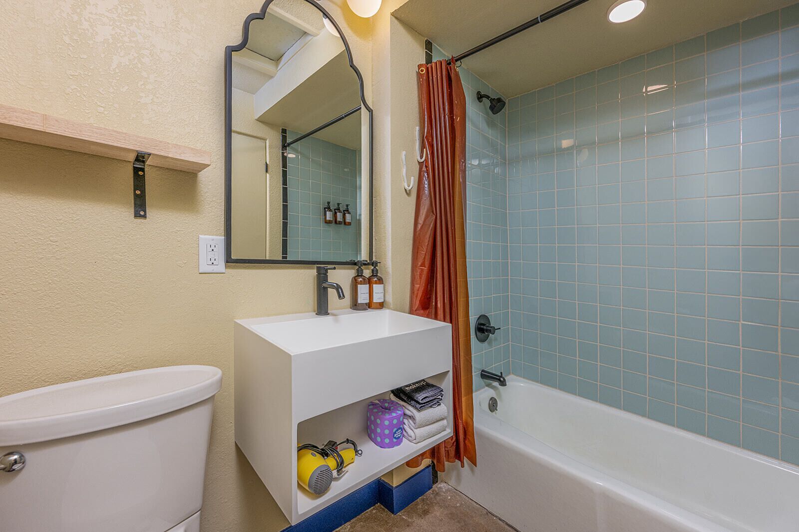 Une salle de bain avec un lavabo blanc, un miroir et des toilettes. On y trouve un rideau de douche et une baignoire dont les carreaux sont bleus.
