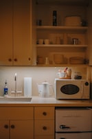 Una cocina con armarios blancos, microondas, hervidor y lavabo con dispensador de jabón.