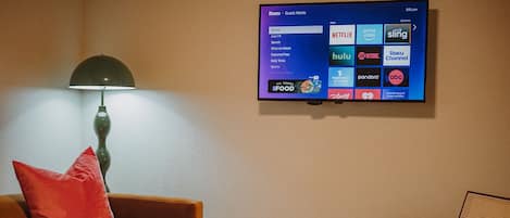 A hotel room with a flat-screen TV displaying the Roku interface, a brown sofa with a red pillow, a small table with a vase, and a chair.