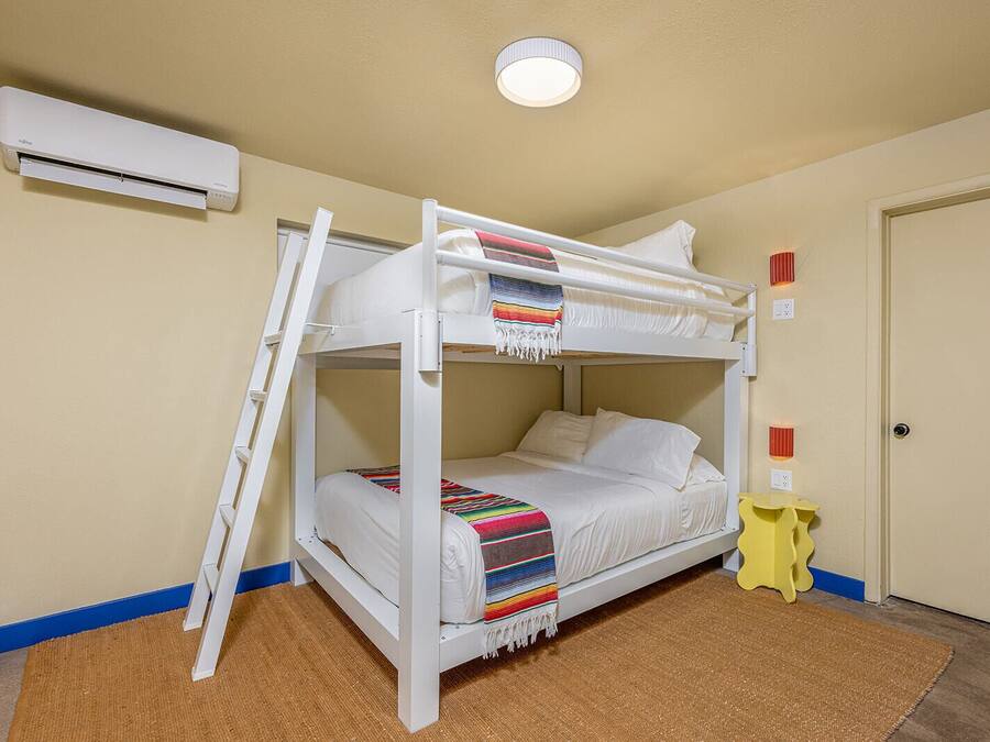 A bunk bed with a ladder, white bedding, a colorful blanket, a small yellow stool, and an air conditioning unit on the wall.
