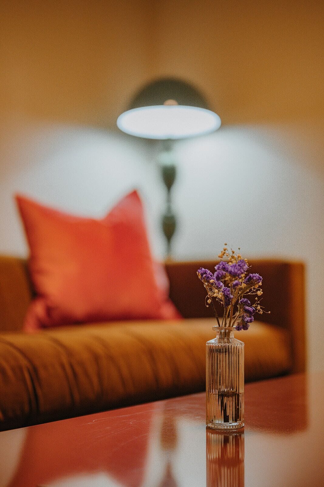 A clear glass vase with dried purple flowers on a wooden table in a room with a brown sofa and a red pillow.