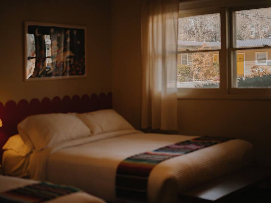A bedroom with a bed, a nightstand with a lamp, a window with a view of a building, and a painting on the wall.