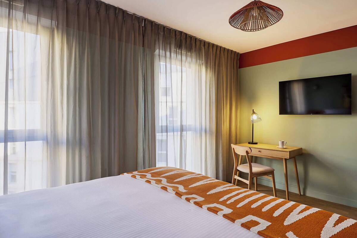 Une chambre d’hôtel équipée d’un lit, d’un bureau, d’une chaise, d’une télévision et d’une grande fenêtre avec des rideaux.