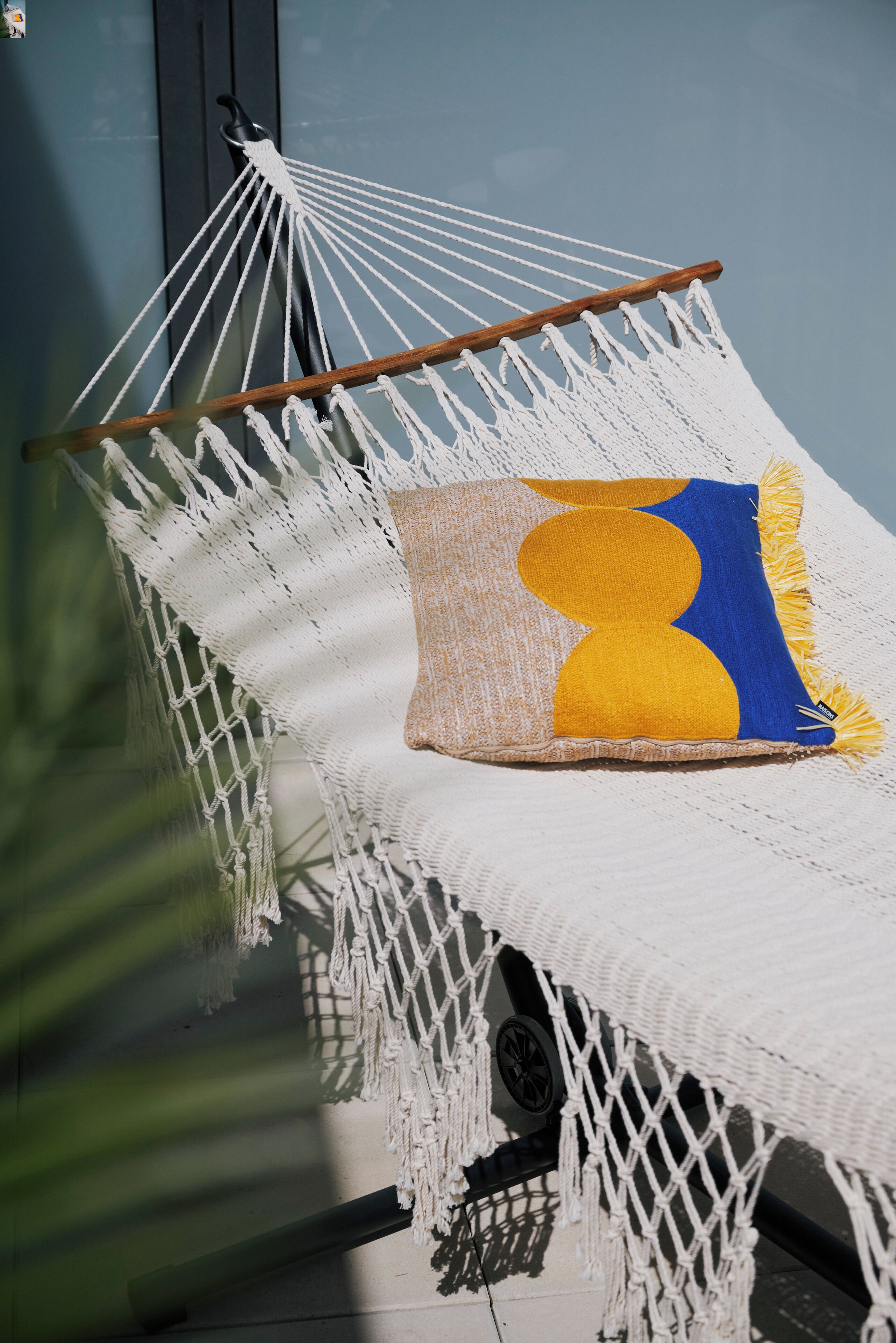 A hammock with macrame netting, a cushion with a geometric pattern, and a wooden support.