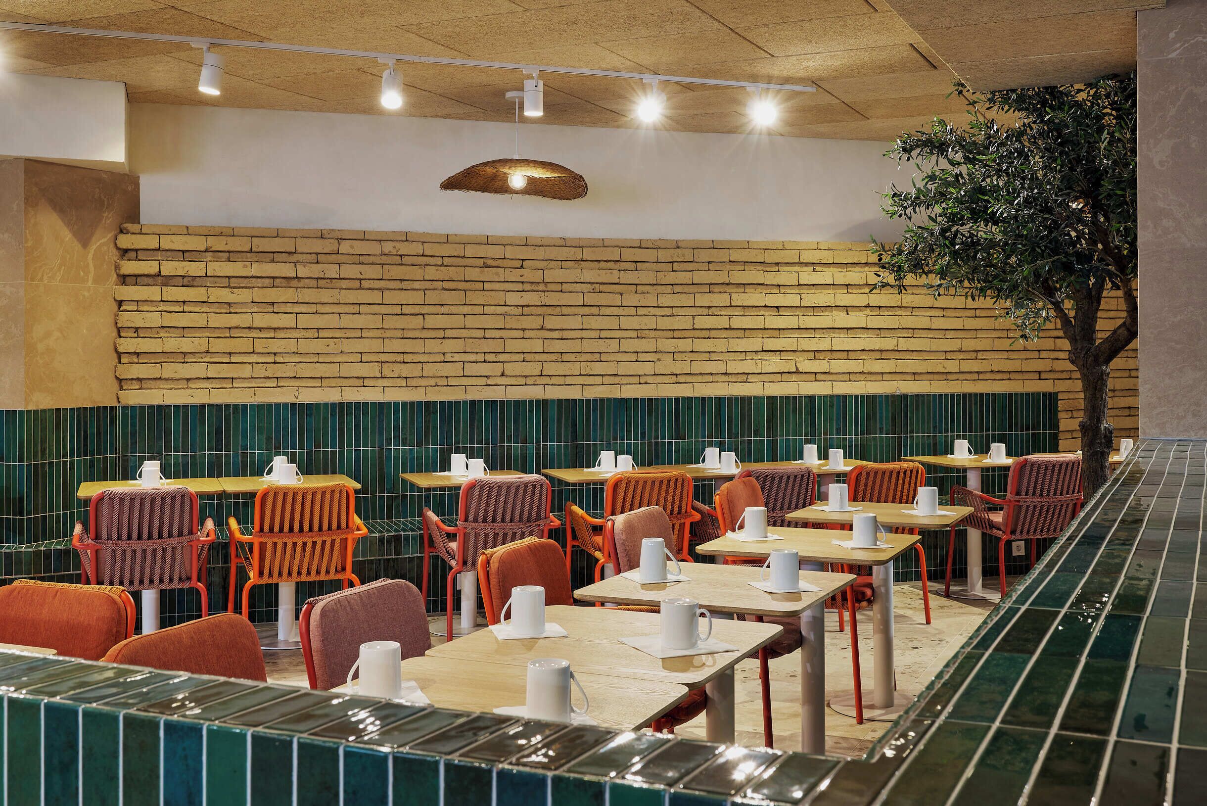 Un restaurant aux murs de briques, avec des banquettes aux carreaux verts et des chaises orange. Les tables sont décorées de sets de table blancs et de tasses.