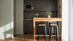 A modern kitchen with dark cabinets, a microwave, a wooden table, and two black bar stools. - Hôtel LIGO by HappyCulture (Marseille)