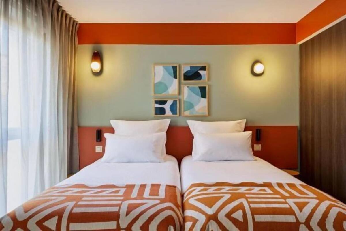 Une chambre d’hôtel avec deux lits, une tête de lit décorée et une fenêtre avec des rideaux.