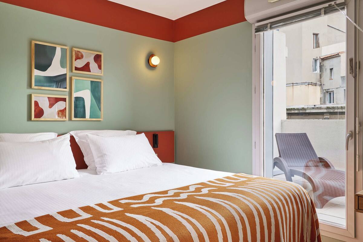 Une chambre d’hôtel avec un lit, un balcon avec une chaise et des œuvres d’art abstraites accrochées au mur.