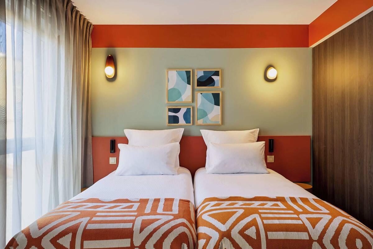 Une chambre d’hôtel avec deux lits, une grande fenêtre avec des rideaux et des lampes fixées au mur.