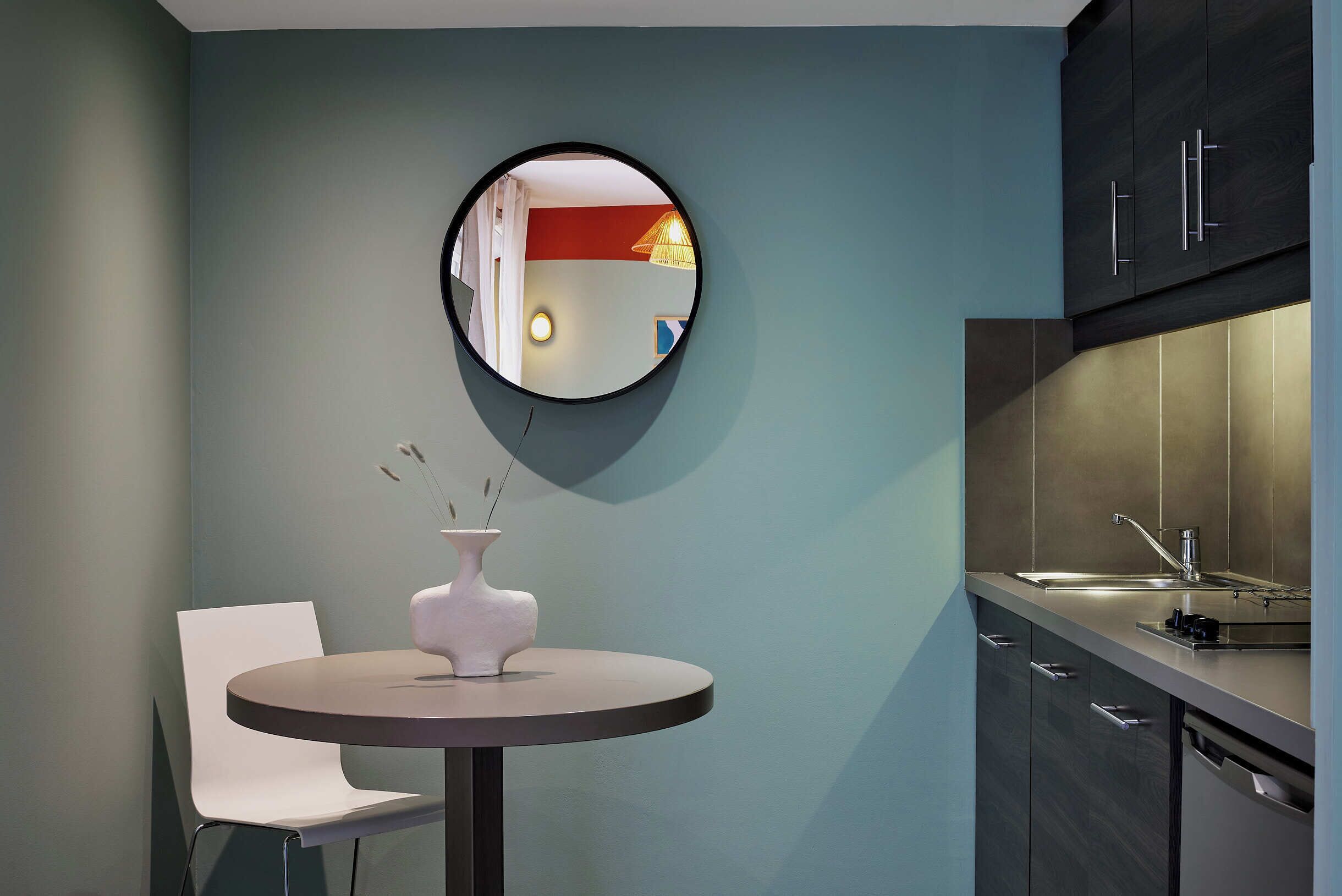 Une cuisine moderne avec des meubles de couleur sombre, un miroir rond, un vase blanc et une petite table avec une chaise.