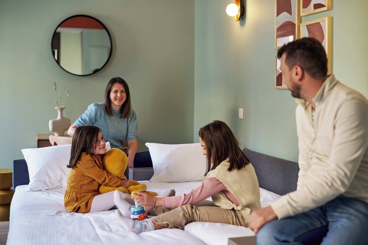 Une famille de quatre personnes est allongée sur un lit, devant un miroir et avec des œuvres d’art accrochées au mur, dans une pièce aux couleurs neutres.
