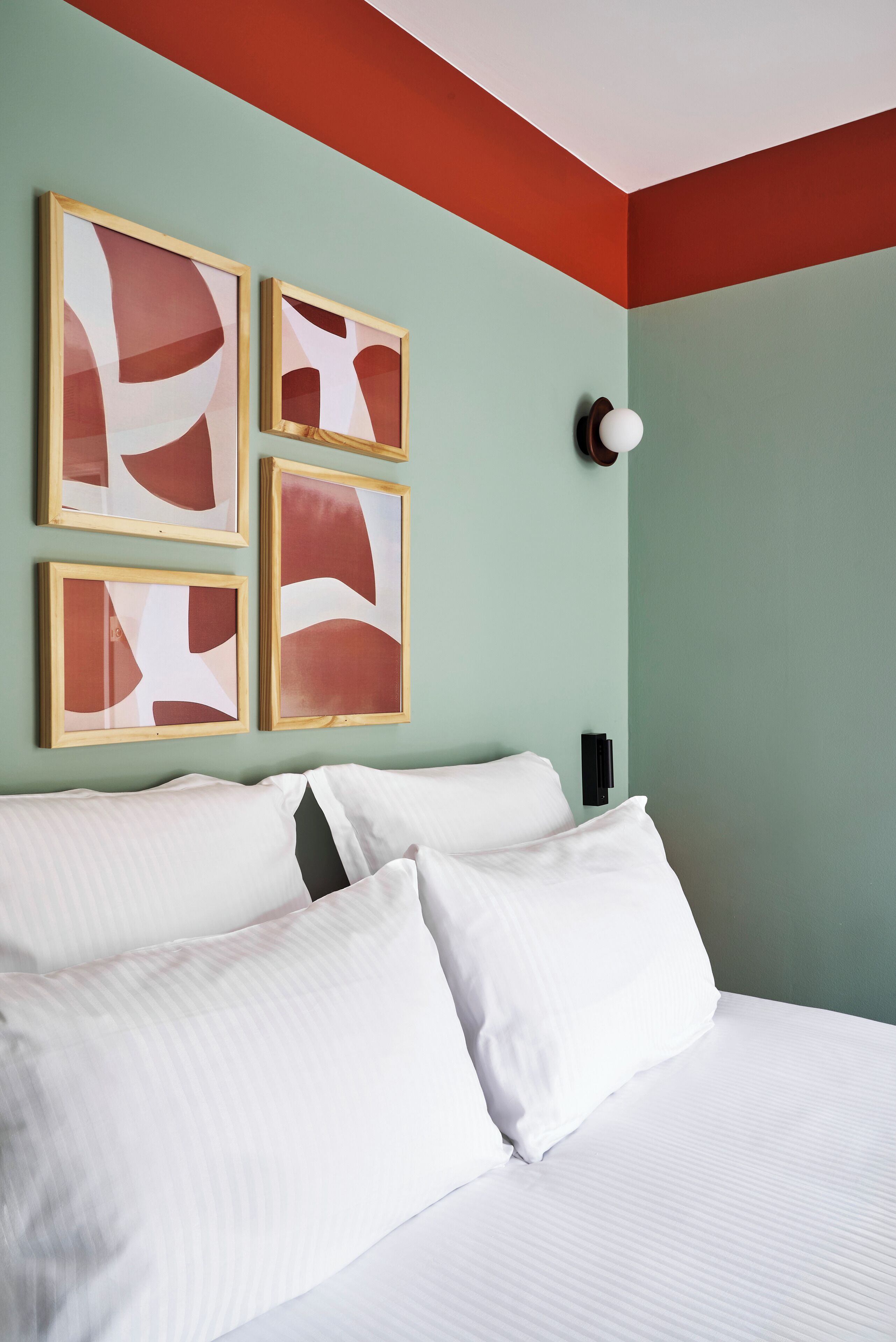 A neatly made bed with white linens and two pillows, a wall-mounted light fixture, and four framed abstract paintings on the wall.