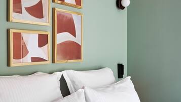 A neatly made bed with white linens and two pillows, a wall-mounted light fixture, and four framed abstract paintings on the wall.