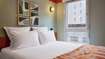 A hotel room with a bed, a window with a view of buildings, and abstract art on the wall.