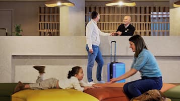 Eine Lobby mit einer Rezeption, einem Kind, das auf einem Pouf spielt, und einer Frau, die sich mit einem Mann unterhält.