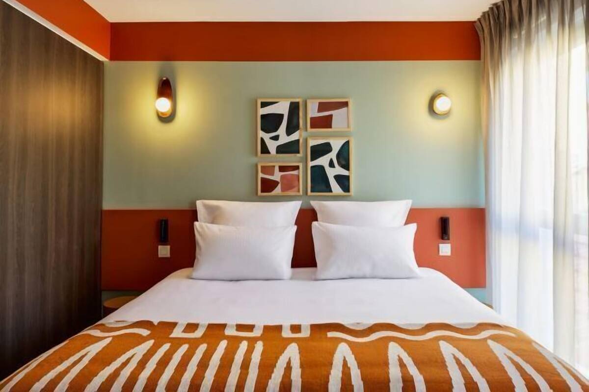 Une chambre d’hôtel avec un lit, deux lampes fixées au mur et une fenêtre avec des rideaux.