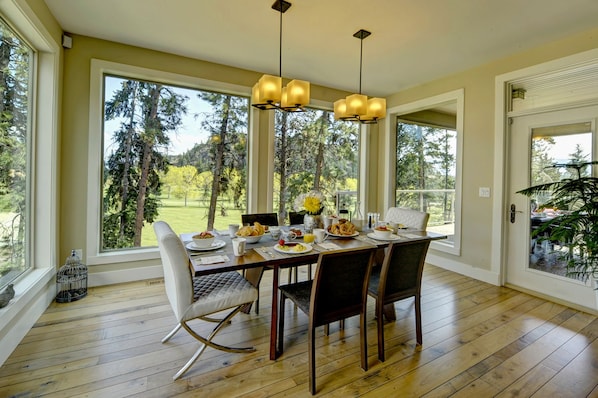 Un comedor con una mesa de madera, sillas blancas con tapicería y vistas a un bosque a través de amplios ventanales.