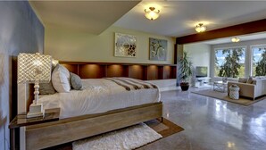 Ein geräumiges Schlafzimmer mit einem großen Bett, einem Nachttisch, einer Lampe und Blick ins Freie durch große Fenster.