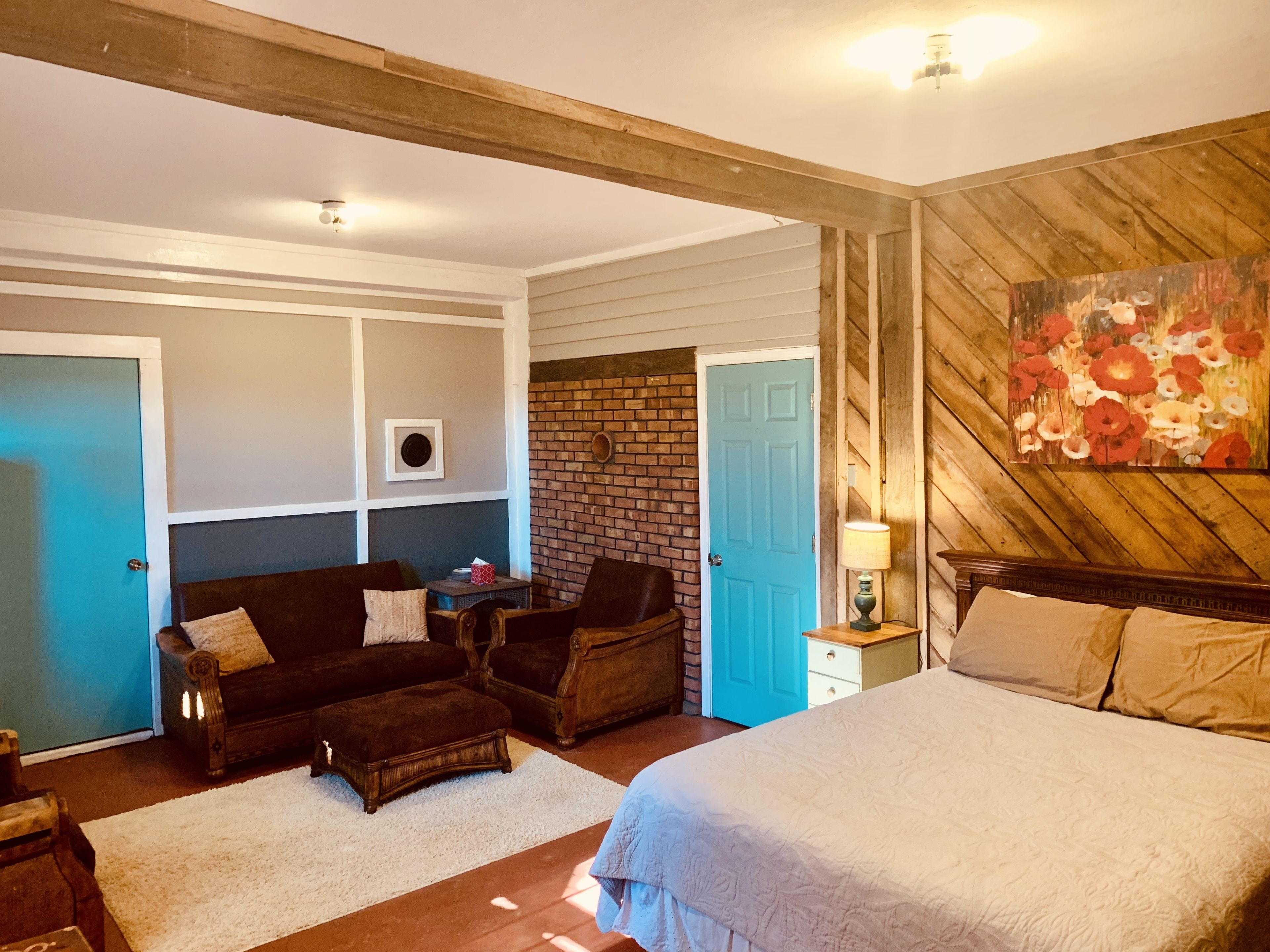 A bedroom with a bed, two armchairs, a small table, a blue door, and a brick wall.