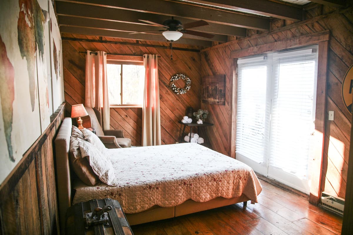 A wooden cabin bedroom with a bed, a nightstand, a dresser, a ceiling fan, and a window with curtains.