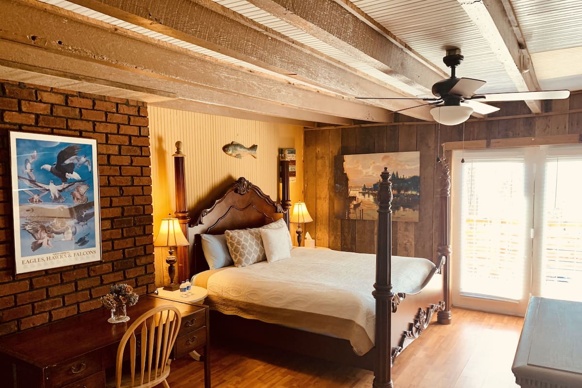 A bedroom with a four-poster bed, wooden walls, a brick accent wall, a ceiling fan, and a large window.