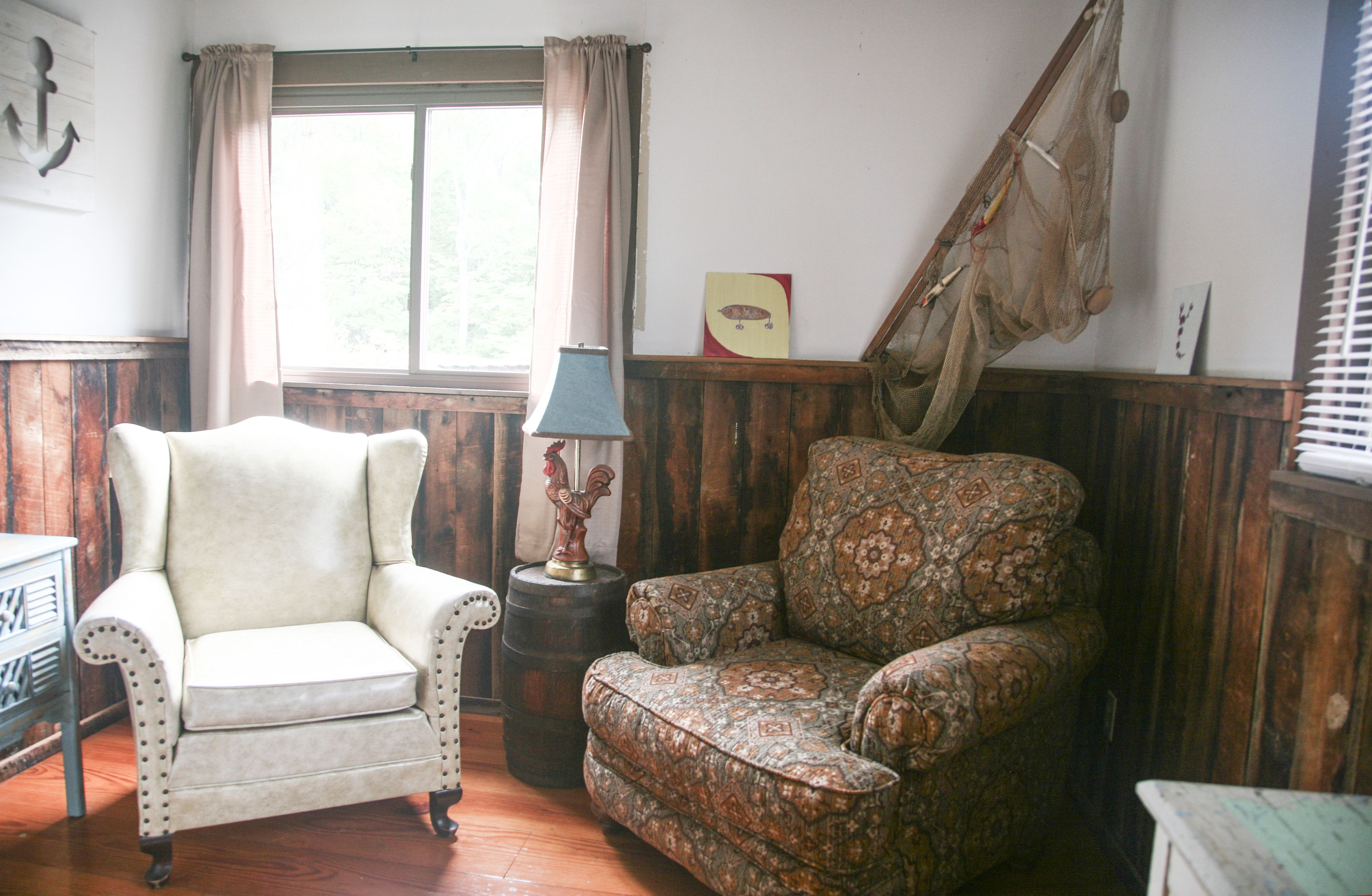 A room with a wooden paneled wall, a white armchair, a patterned armchair, a small table with a lamp, and a window with curtains.