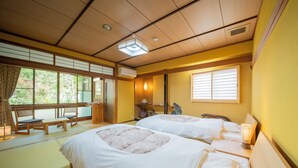 A traditional Japanese-style room with tatami flooring, two beds with white bedding, a wooden bench, and a window with blinds. - Yanagiso (Toyooka)