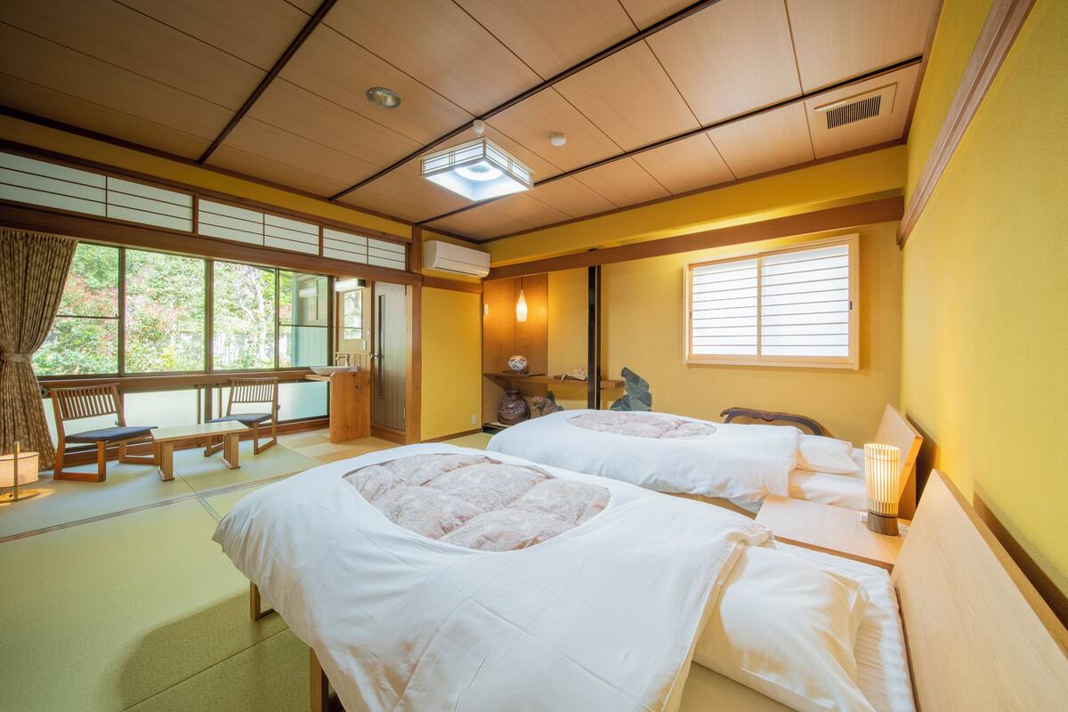 A traditional Japanese-style room with tatami flooring, two beds with white bedding, a wooden bench, and a window with blinds.