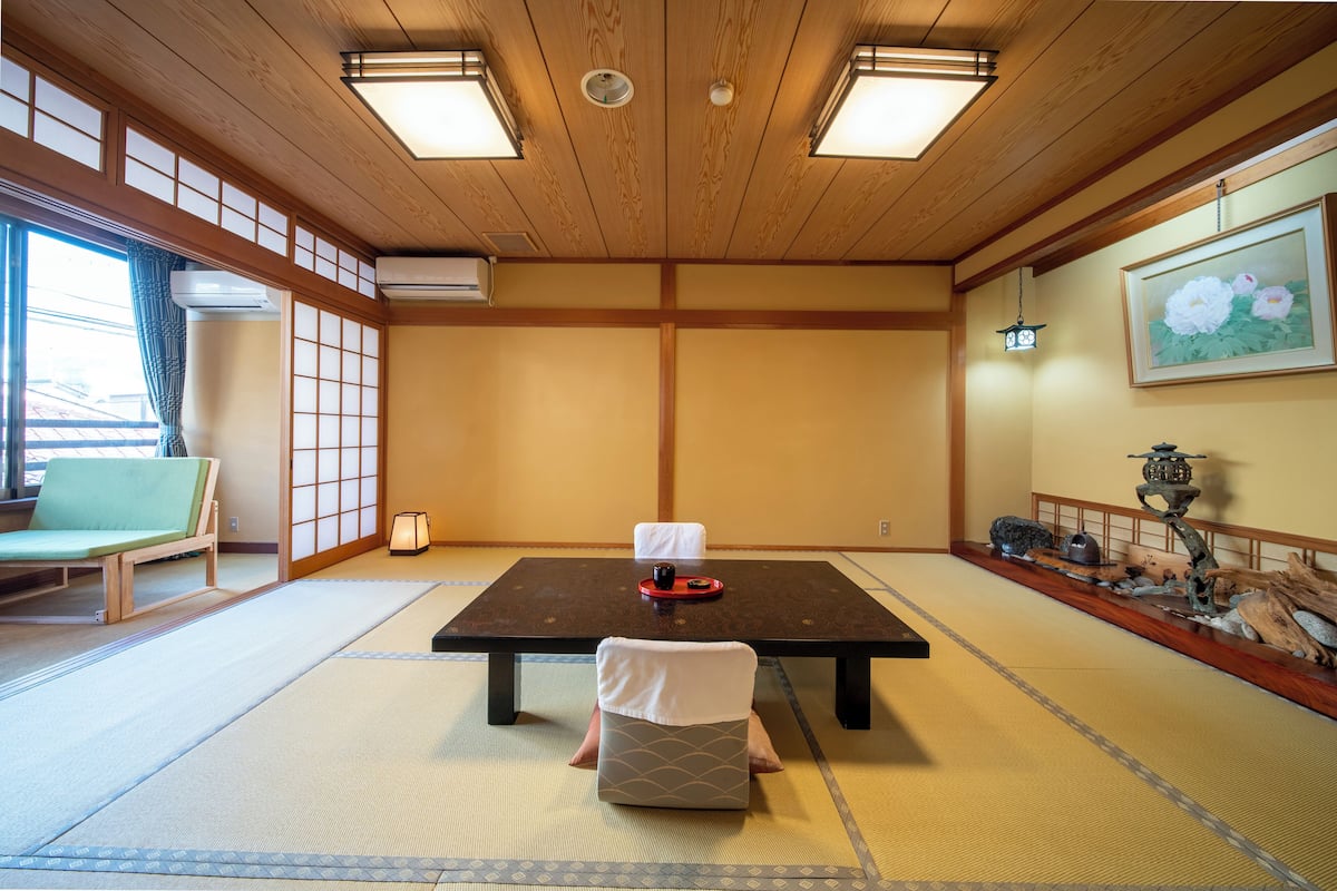 A traditional Japanese room with tatami flooring, a low table with a cushion, a green chair, and a decorative lantern.