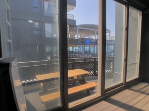 Uitzicht vanaf een balkon met een houten tafel en stoelen, een glazen deur, en een gebouw met een caféborduur in de verte.