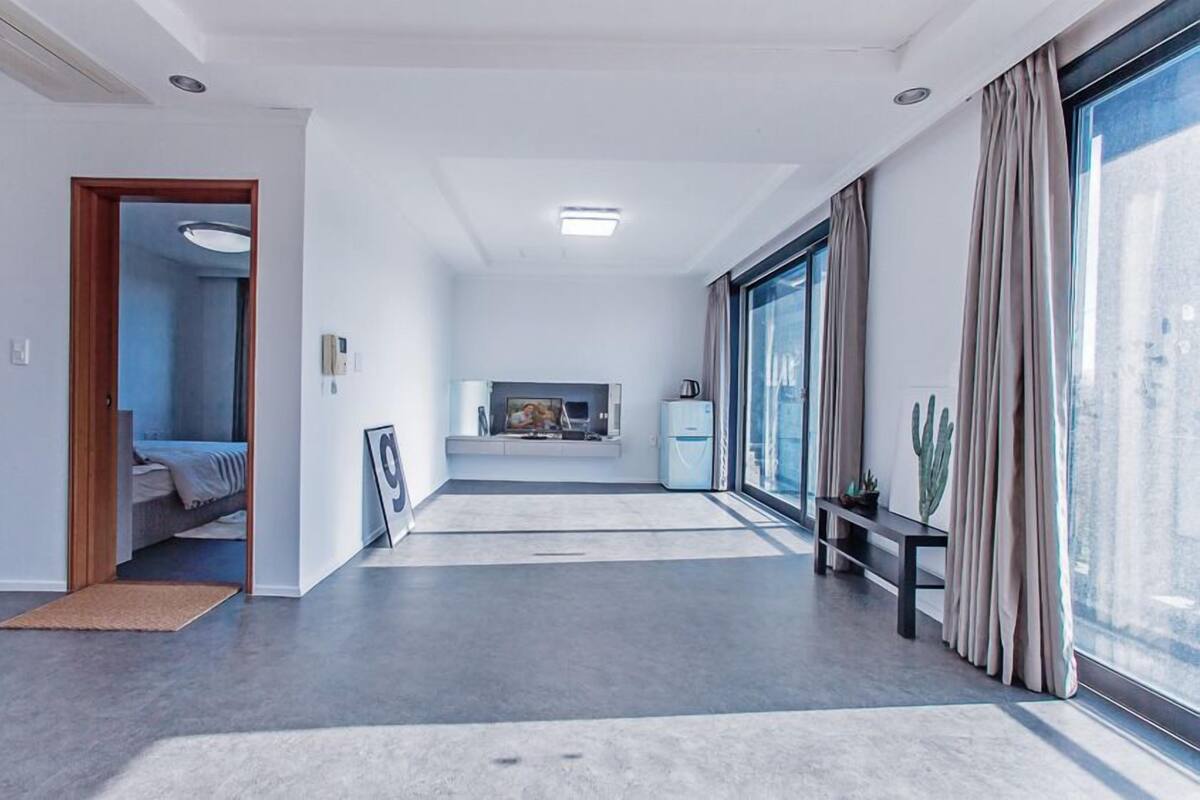 A modern hotel room with a large window, a bed, a TV, and a small kitchenette.