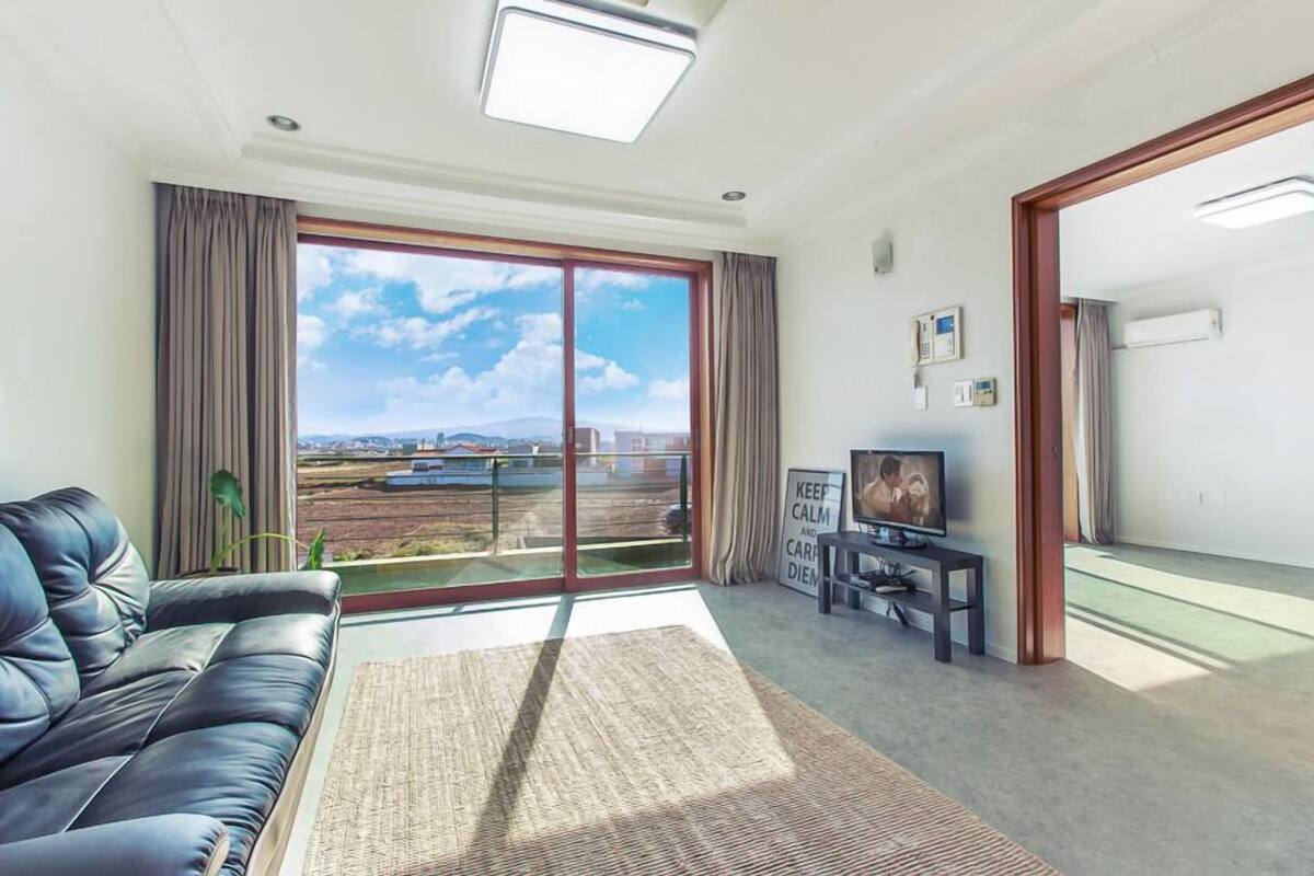 A living room with a black leather sofa, a flat-screen TV, and a large sliding glass door leading to a balcony with a view of the cityscape.