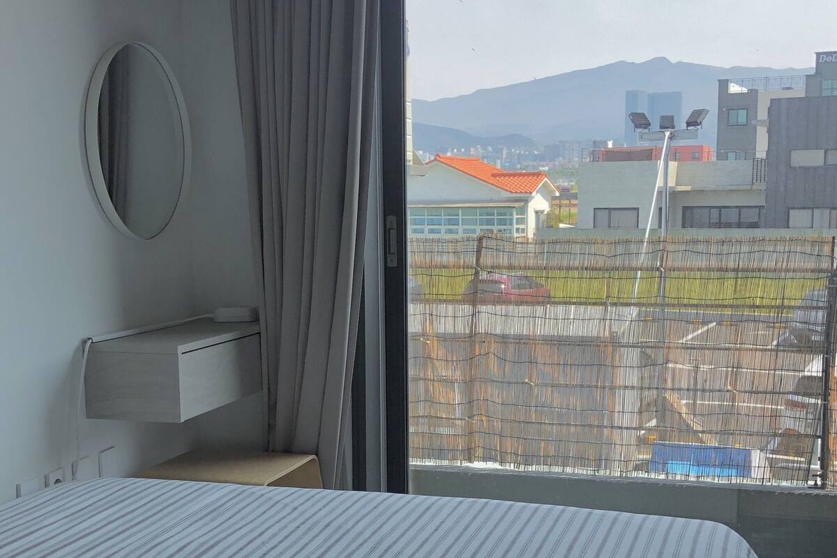 A hotel room with a bed, a bedside table, a mirror, and a window with a view of buildings and a fence.