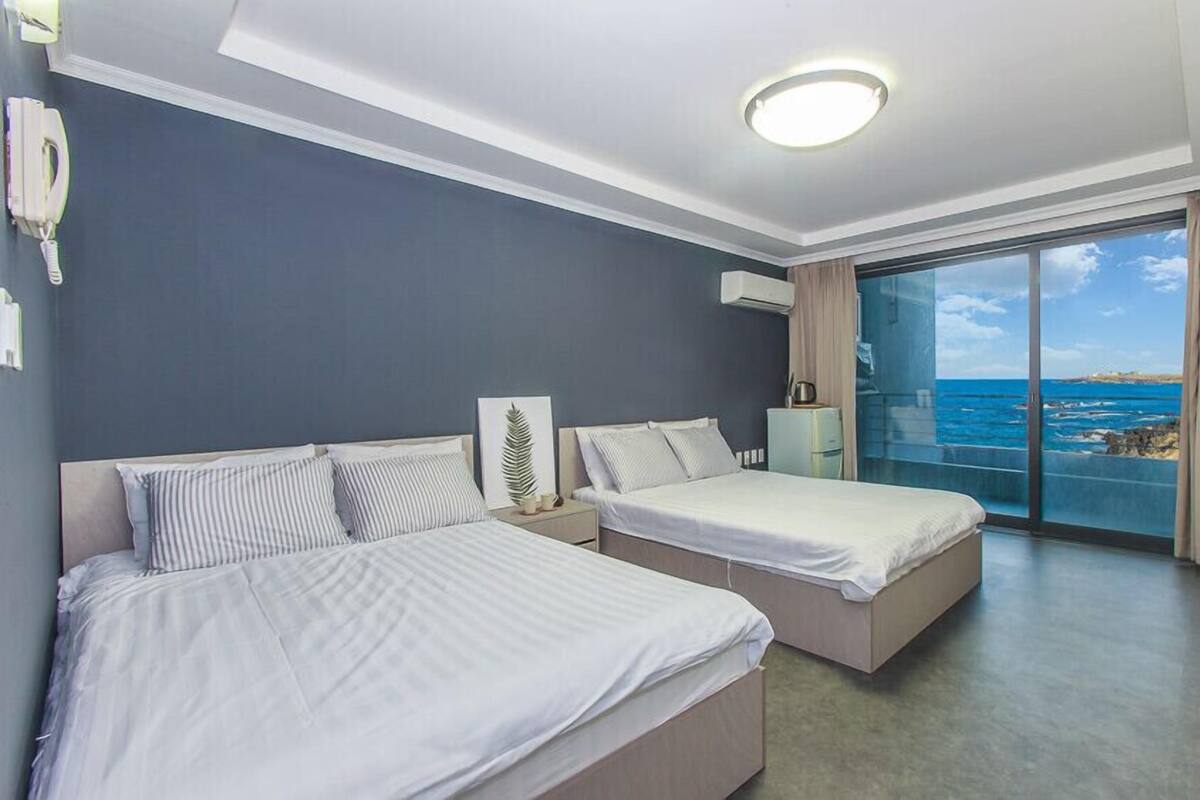 A hotel room with two beds, a large window with a view of the sea, a ceiling light, and a phone on the wall.