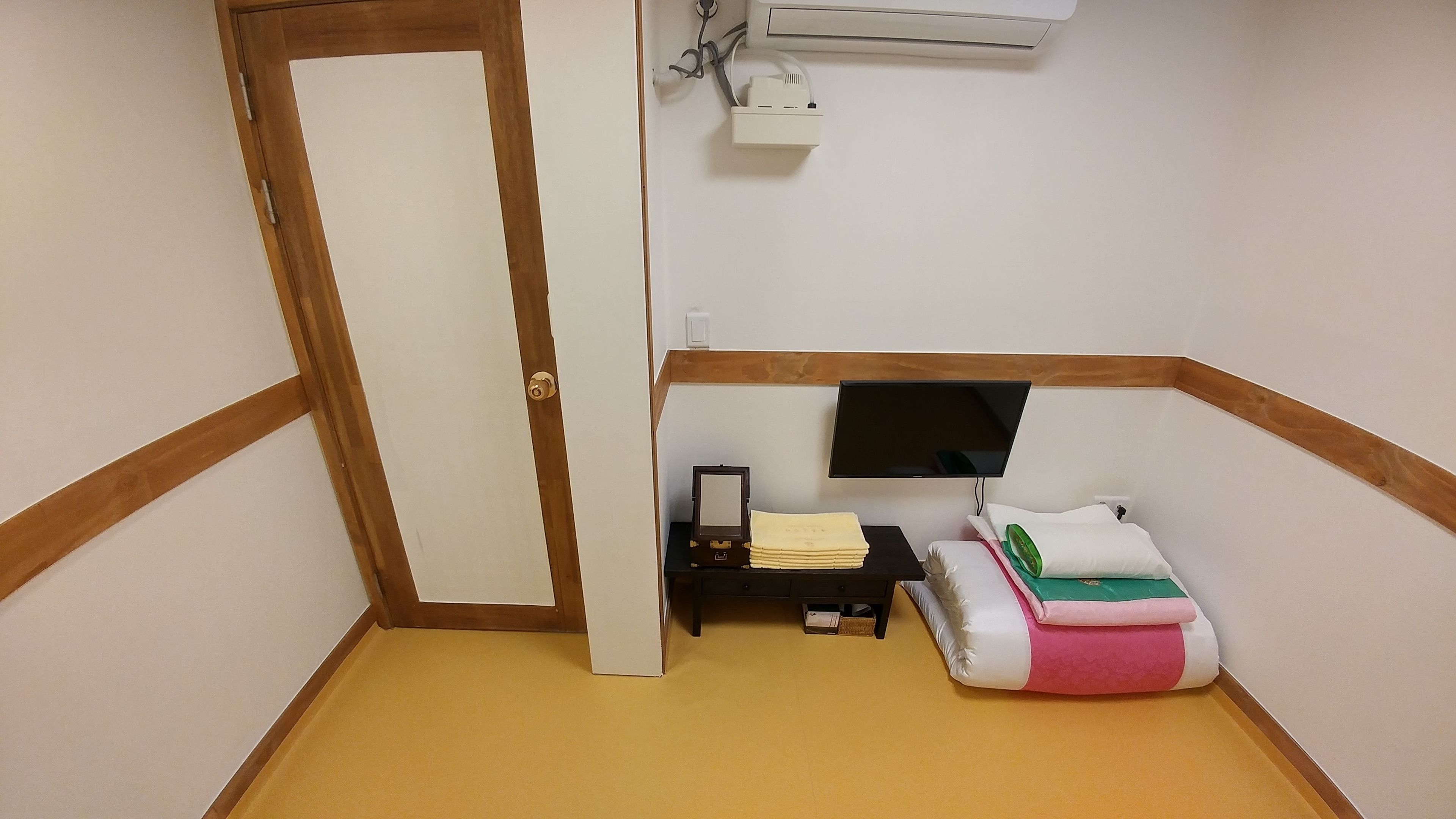 Una habitación pequeña y ordenada con una puerta, un equipo de aire acondicionado, un televisor de pantalla plana montado en la pared, una pequeña mesa con una pila de documentos y un cojín colorido en el suelo.