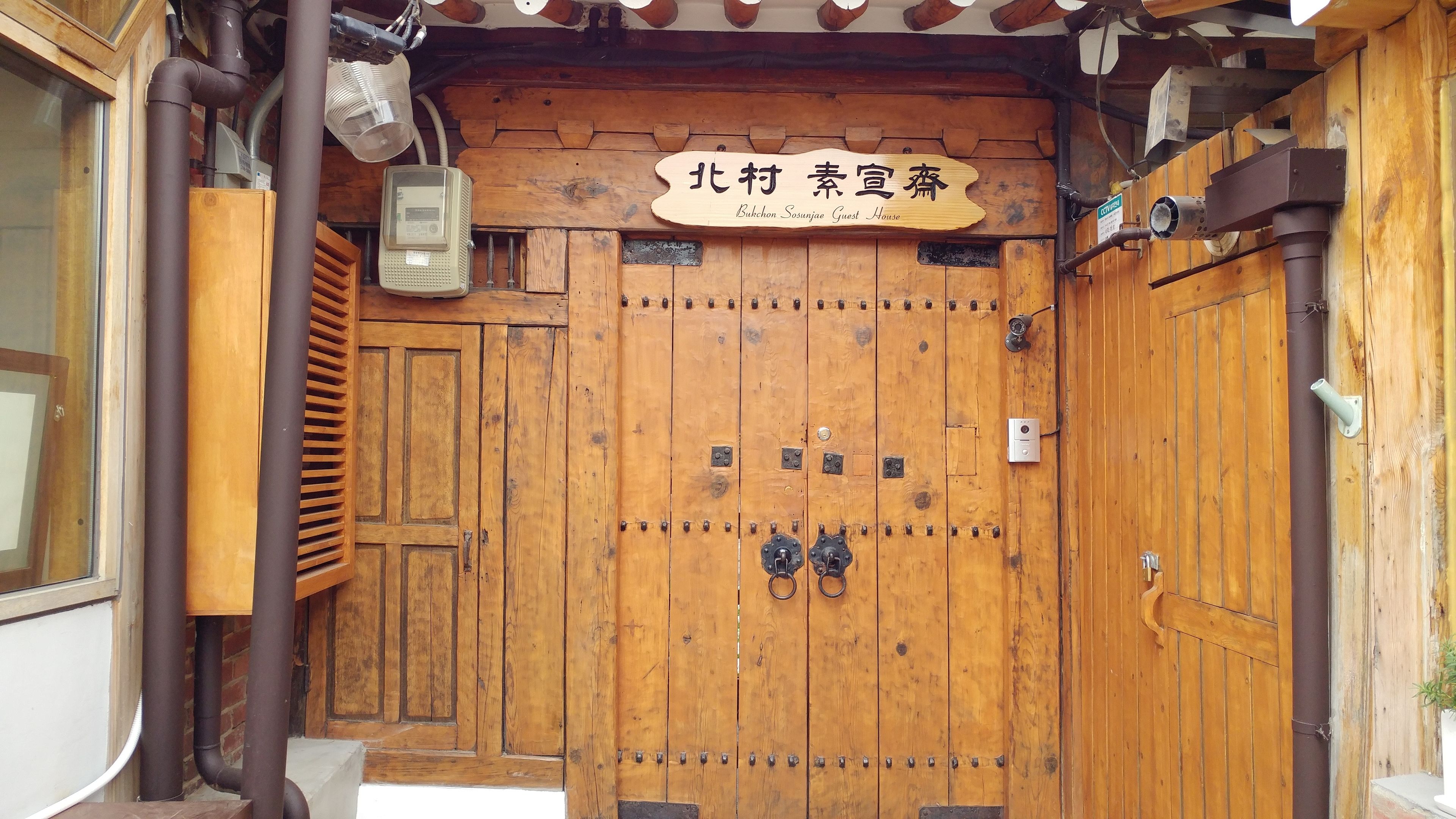 A wooden door with a sign above it, a metal vent, and a small electronic device attached to the wall.